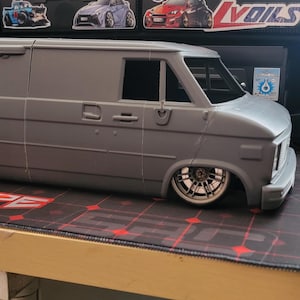 May include: A gray model van with lowered suspension and custom wheels. The van is a detailed replica, showcasing a classic design. It sits on a patterned surface, suggesting a display or hobbyist setting. The vehicle's body is a solid gray color.