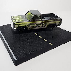 May include: A black and silver toy pickup truck with yellow flames painted on the side, displayed on a black and white road diorama.