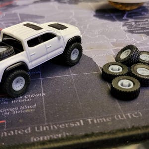 May include: A white toy pickup truck with black wheels and a spare tire in the bed. Several additional black tires with gray rims are next to the truck. The truck is on a map-themed surface.
