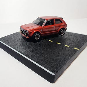 May include: A red toy car with black accents and a black spoiler is displayed on a black and white miniature road surface.