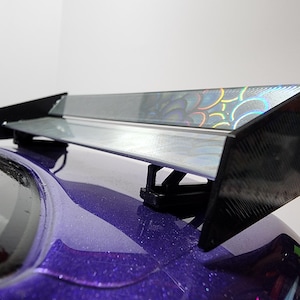 May include: A black and silver car spoiler with a holographic design. The spoiler is mounted on the back of a purple car.