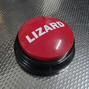 May include: A large, red button with the word "LIZARD" in white, bold letters. The button has a black base and sits on a textured, metallic surface. The button is round and has a glossy finish.