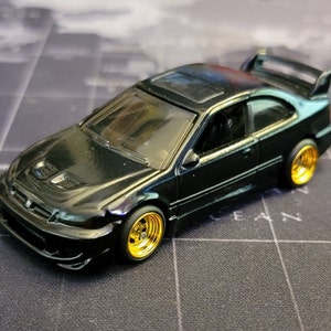 May include: A black die-cast model car with gold-colored rims and a rear spoiler. The miniature vehicle has a sunroof and a hood scoop. The car is set against a dark background.