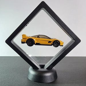 May include: A yellow toy sports car with black wheels and accents is displayed inside a black, diamond-shaped frame. The frame is mounted on a black base, creating a floating effect. The car has a sleek design, visible through the clear front of the frame.