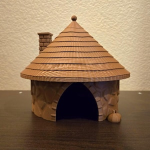 May include: A brown 3D printed miniature house with a conical roof, a textured stone-like wall pattern, a chimney on the left side, and a large arched doorway. A small pumpkin-shaped object sits on the ground to the right of the entrance.