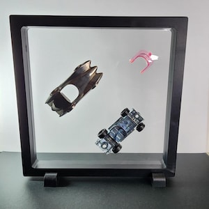 Può includere: Una cornice nera a scatola contenente tre parti di auto giocattolo. Le parti includono un'auto argentata in stile Batmobile, un pezzo rosa e bianco e un'auto blu e nera con ruote. La cornice è esposta su una superficie scura.
