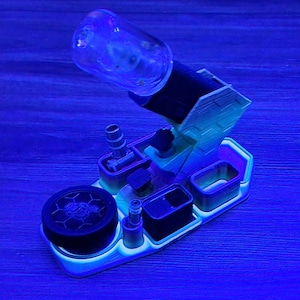 May include: A unique, multi-component device with a clear glass bottle, black cylindrical section, and a light green base. The base has various compartments and a circular black piece with a honeycomb pattern. The device is illuminated with a blue light.