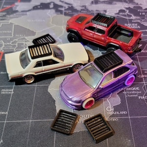 May include: Three toy cars with black roof racks. The cars are a white sedan, a purple hatchback, and a red pickup truck. The cars are on a map with a black background.