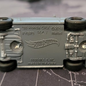 May include: Underside view of a gray Hot Wheels die-cast model car. The text on the car reads "'88 Honda CRX &copy;2018 FYB70 M47 Mattel". The car has black wheels and is made in Malaysia.