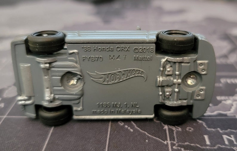May include: Underside view of a gray Hot Wheels die-cast model car. The text on the car reads "'88 Honda CRX &copy;2018 FYB70 M47 Mattel". The car has black wheels and is made in Malaysia.