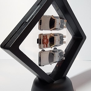 May include: Three silver toy cars, possibly DeLorean time machines, are displayed in a black diamond-shaped frame with a black base. The cars are suspended in the frame, giving the illusion of floating.