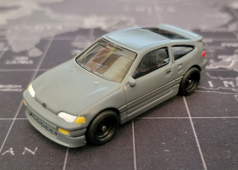 May include: A gray die-cast model car with black wheels. The miniature vehicle has a sleek design, featuring a detailed front end with yellow and white accents. The car is a miniature replica of a classic sports car.