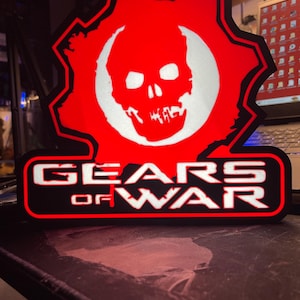Puede incluir: Logotipo rojo y negro de Gears of War con una calavera blanca en el centro. El texto "GEARS OF WAR" está en letras blancas. El logotipo está iluminado, creando una pantalla brillante.