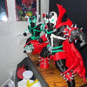 May include: Two Spawn action figures in a dynamic pose. The foreground figure features a black suit with white accents, a red cape, and silver spikes on the gauntlets and boots. The background figure has a black suit with green accents and a white cape.