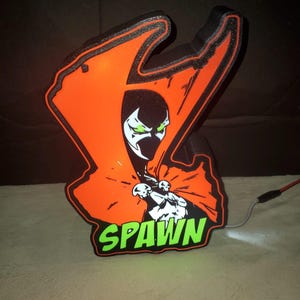 May include: An orange and black Spawn-themed lamp featuring the character's image and the word "SPAWN" in green. The lamp has a black outline and a cord for power.