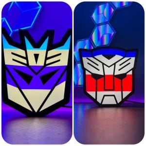 May include: Two Transformers robot logos, one in purple, white, and blue, and the other in red, white, and blue, are illuminated with colorful lights. The logos are displayed on a dark background.