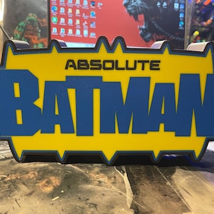 May include: A yellow and blue 3D printed sign with the text "ABSOLUTE BATMAN" in black letters.