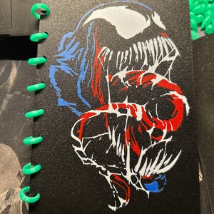 May include: A black notebook with bright green plastic binding discs. The cover features a graphic illustration of a character with a wide, toothy grin, rendered in white, red, and blue lines against the textured black background.