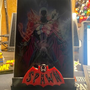 May include: A black and red 3D printed stand with a red bat-shaped logo that says "SPAWN" in black letters. The stand is holding a clear plastic frame with a comic book image of Spawn, a superhero with a red cape and a skull-like helmet.