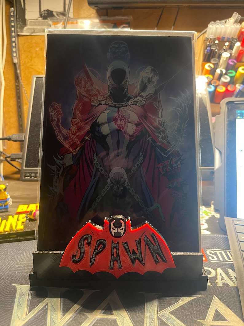 May include: A black and red 3D printed stand with a red bat-shaped logo that says "SPAWN" in black letters. The stand is holding a clear plastic frame with a comic book image of Spawn, a superhero with a red cape and a skull-like helmet.