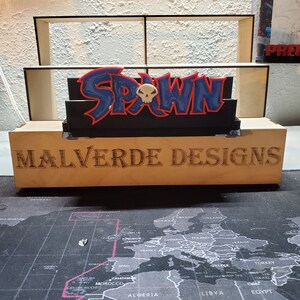 May include: A wooden shelf unit with a 3D printed Spawn logo. The logo is blue, red, and black with a white skull. The shelf unit has the words "Malverde Designs" printed on the front.