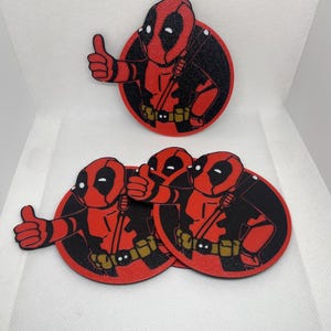 May include: Set of four round coasters featuring a cartoon image of Deadpool giving a thumbs up. The coasters are red with a black outline and a gold belt.