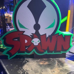 May include: A 3D printed figurine of the Spawn character from the comic book series. The figurine is green and red with a white skull and the word "Spawn" in bold letters.