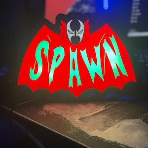 May include: A red and black light-up sign with the word "SPAWN" in green letters. The sign features a cartoon image of Spawn, a comic book character, with a red cape and a black mask.