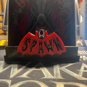 May include: A red and black 3D printed figurine of Spawn, a comic book character. The figurine is shaped like a bat with the word "Spawn" written in black letters across the wings. The figurine is sitting on a black base.