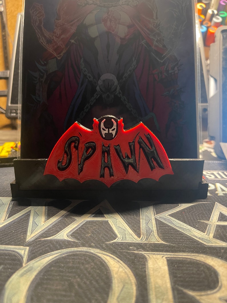 May include: A red and black 3D printed figurine of Spawn, a comic book character. The figurine is shaped like a bat with the word "Spawn" written in black letters across the wings. The figurine is sitting on a black base.