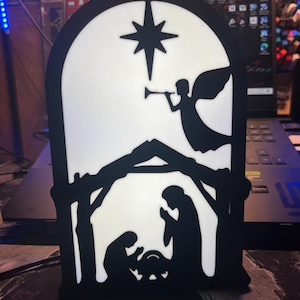 May include: A black silhouette of a nativity scene with a star above the manger. The scene is illuminated from behind, creating a glowing effect.