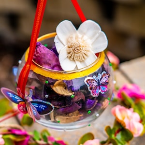Pode incluir: Um frasco de vidro com uma borda dourada e uma fita vermelha pendurada no topo. O frasco está cheio de potpourri e uma flor branca. Há duas borboletas no frasco, uma azul e uma rosa.