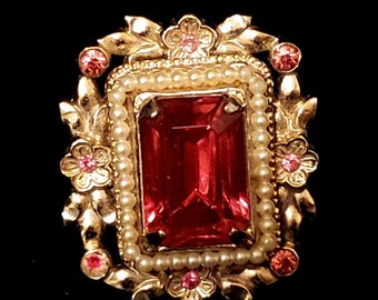 Könnte beinhalten: Eine goldfarbene Brosche mit einem großen, rechteckigen roten Stein, der von einem Rand aus falschen Perlen umgeben ist. Die Brosche ist mit kleinen rosa Blüten und Blättern verziert.