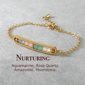 Könnte beinhalten: Goldkette Armband mit einem Bar-Anhänger mit Aquamarin, Rosenquarz, Amazonit und Mondstein Perlen. Das Armband ist mit "Nurturing" beschriftet.