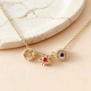 May include: A gold-colored necklace featuring three flower pendants. The flowers have a textured design and are adorned with a clear, red, and blue gemstone center. The necklace is displayed on a marble-patterned surface.
