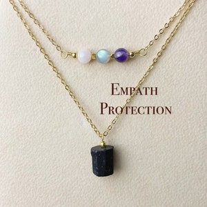 May include: Gold chain necklace with a black stone pendant and a smaller chain with three beads: pink, blue, and purple. The necklace has the text "Empath Protection" on the card.