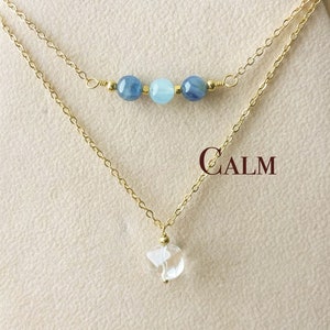Può includere: Una collana con catena dorata con un ciondolo di cristallo trasparente e una fila di perle di pietre blu. La collana è esposta su una carta con la scritta "CALM".