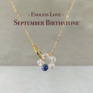 May include: A gold chain necklace with a pendant made of clear and blue beads. The pendant is in the shape of a circle with a blue bead in the center. The necklace has the text "Endless Love - September Birthstone" on it.