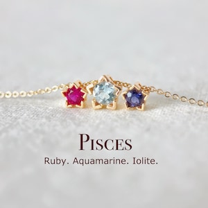 May include: A delicate gold necklace featuring three star-shaped pendants with gemstones. The gemstones are ruby, aquamarine, and iolite. The word "Pisces" is displayed below the necklace.