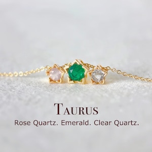 May include: A gold necklace with three star-shaped pendants, each holding a gemstone: rose quartz, emerald, and clear quartz. The word "TAURUS" is displayed below the necklace.