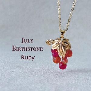 May include: Gold necklace with a grape cluster pendant made of red and orange gemstones. The pendant features a gold leaf design. The text "JULY BIRTHSTONE Ruby" is written below the pendant.
