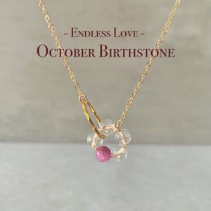 May include: A gold chain necklace with a pink and clear crystal pendant. The pendant is a circle of clear crystals with a pink crystal in the center. The text "Endless Love - October Birthstone" is written above the pendant.