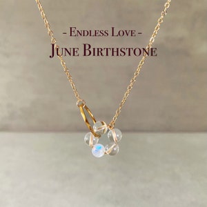 May include: A gold chain necklace with a charm made of clear round beads and a gold ring. The charm is shaped like a flower. The necklace has the text "Endless Love - June Birthstone" on it.