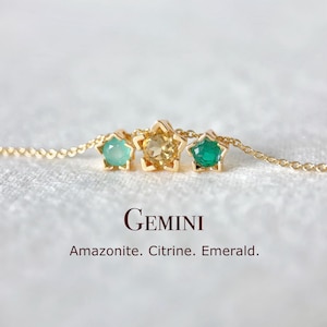 Könnte beinhalten: Eine goldene Halskette mit drei sternförmigen Anhängern, die jeweils einen Edelstein enthalten: Amazonit, Citrin und Smaragd. Das Wort "GEMINI" und die Edelsteinnamen sind darunter angezeigt.