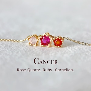Peut inclure: Un collier en or orné de trois pendentifs en forme d'étoile sertis de pierres précieuses : quartz rose, rubis et cornaline. Le mot "CANCER" et les noms des pierres précieuses sont affichés en dessous.