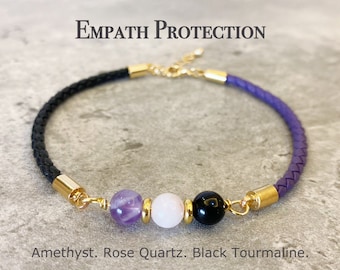 May include: A purple, black, and white braided leather bracelet with gold accents. The bracelet features three gemstone beads: amethyst, rose quartz, and black tourmaline. The text "Empath Protection" is written above the bracelet.