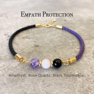 May include: A purple, black, and white braided leather bracelet with gold accents. The bracelet features three gemstone beads: amethyst, rose quartz, and black tourmaline. The text "Empath Protection" is written above the bracelet.