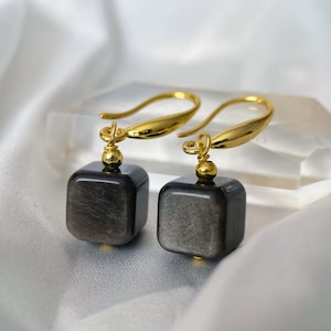 May include: A pair of gold dangle earrings with black square-shaped gemstone drops.