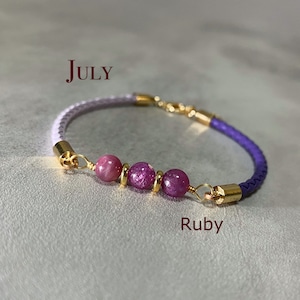 May include: A braided leather bracelet with ruby gemstones and gold details. The bracelet is purple and lavender with three ruby beads. The word "July" is above the bracelet and "Ruby" is below.