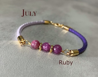 Op de afbeelding: Een gevlochten leren armband met robijn edelstenen en gouden accenten. De armband is paars en lavendel met drie robijn kralen in het midden. Het woord "July" staat boven de armband en "Ruby" eronder.